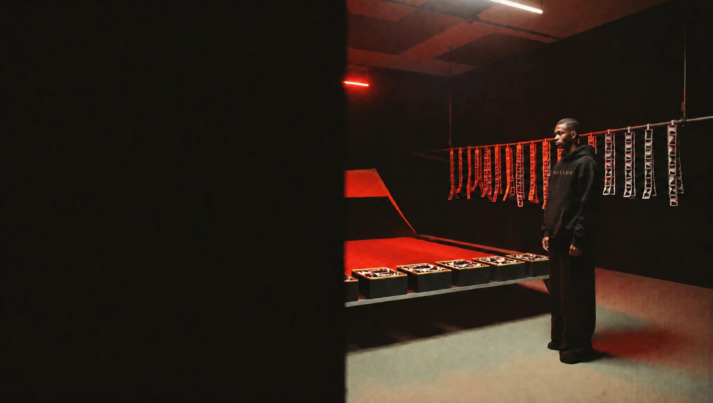 A man in a black heavyweight hoodie stands in a darkroom under red safelight, film strips drying behind him.